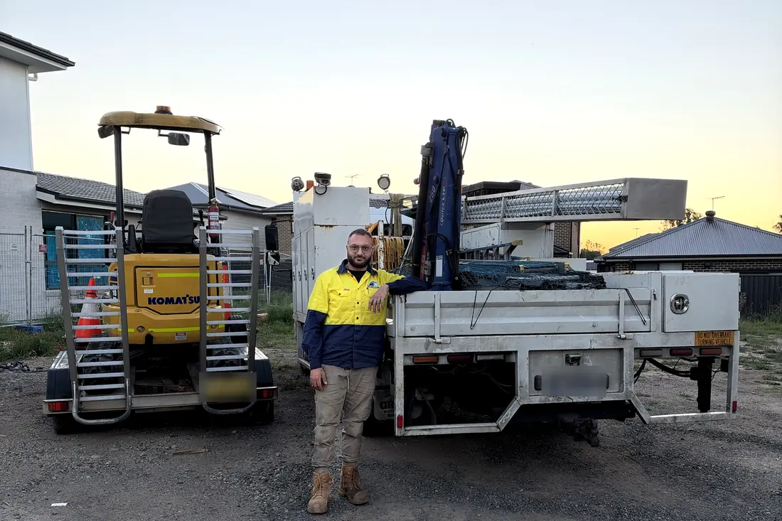 Paul, owner of Monarch Civil & Plumbing, standing between the Komatsu PC18 excavator on its trailer and the crane truck